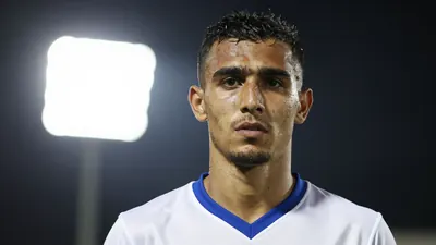 A close-up shot of an anonymous Omani international striker wearing a white and blue football kit, with a floodlight rim...