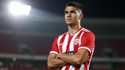 A young, muscular striker in a red and white striped football kit, standing confidently with arms crossed, looking determined