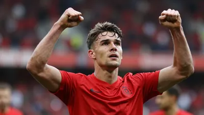 A young Leyton Orient striker in a red shirt celebrating a goal, arms raised, face full of determination, blurred stadium...